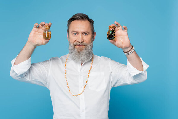 bearded healing guru holding jars with scented oils and looking at camera isolated on blue