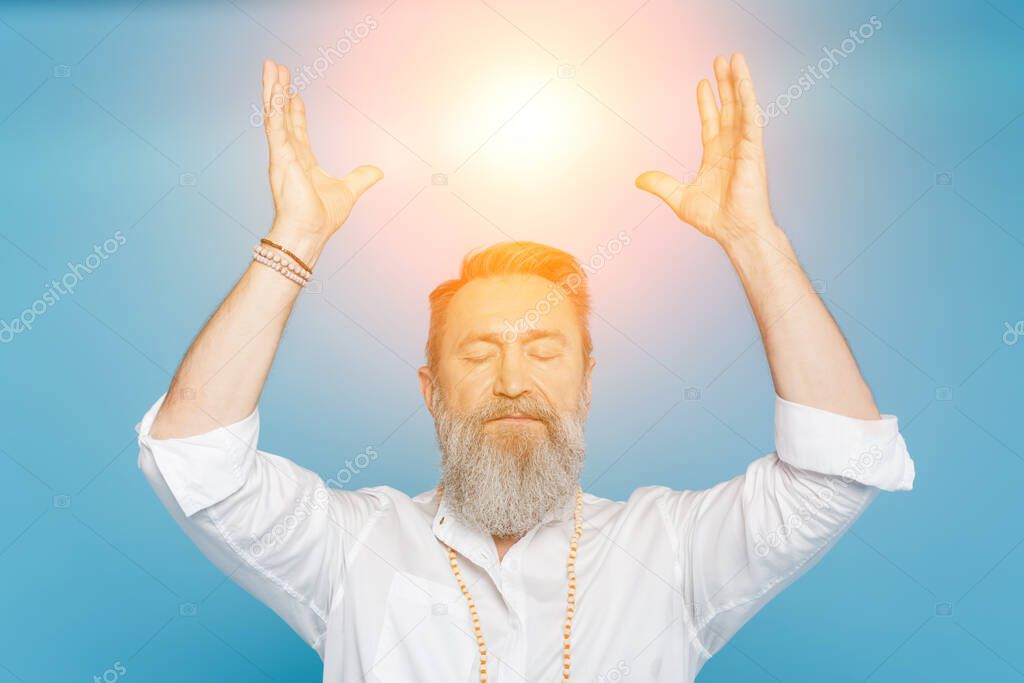 master guru meditating with closed eyes and raised hands near shining aura isolated on blue