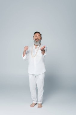 full length of pleased yoga coach in white clothes looking at camera and pointing with hand on grey clipart