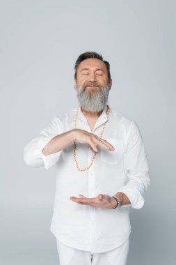 Sakallı guru koçu, gri renkte kapalı gözlerle meditasyon yaparken el kol hareketi yapıyor.