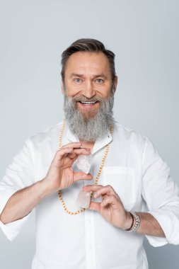 senior spiritual coach holding crystals of selenite while smiling at camera isolated on grey clipart