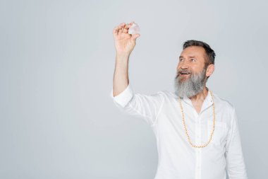 happy guru man with grey beard holding selenite crystal isolated on grey clipart