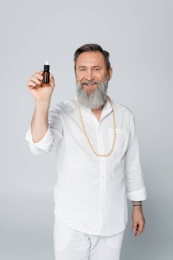 Sakallı şifacı guru, yağ şişesi tutuyor ve gri kameraya gülümsüyor.