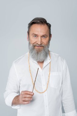 Sakallı şifacı guru aroma çubuğu tutuyor ve gri renkli kameraya bakıyor.