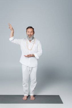 Beyaz elbiseli, sakallı ve yalın ayaklı guru. Gri renkli kameraya bakıyor.