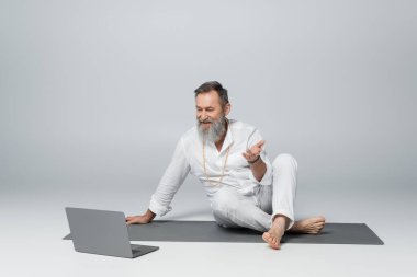 Guru akıl hocası yoga minderinde bacak bacak üstüne atmış oturuyor ve gri dizüstü bilgisayarın yanında konuşuyor.