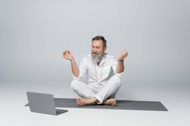 Gri saçlı yoga ustası rahat bir poz veriyor ve gri dizüstü bilgisayarın yanında çenesini gösteriyor.