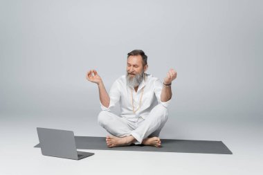 grey haired yoga master sitting in easy pose and showing chin mudra near laptop on grey clipart