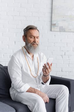 Beyaz tişörtlü ve boncuklu sakallı guru koltukta cep telefonuyla oturuyor.