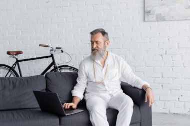 Beyaz elbiseli kıdemli deha koçu evde kanepede otururken dizüstü bilgisayar kullanıyor.