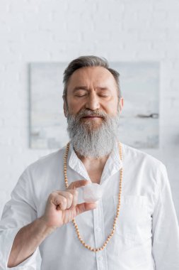 Sakallı guru adam evde gözleri kapalı ve selenit taşıyla meditasyon yapıyor.