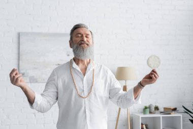 Beyaz gömlekli ve boncuklu sakallı akıl hocası evde gözleri kapalı meditasyon yapıyor.