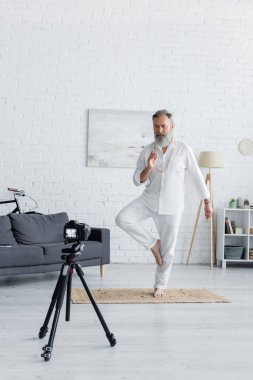 Yaşlı yoga koçu ağaç duruşunu ve dijital kameranın önünde prana Mudra hareketini gösteriyor.