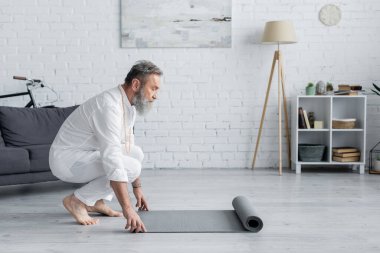 Beyaz elbiseli yaşlı bir adamın evde yoga minderini açarken yan görüntüsü.
