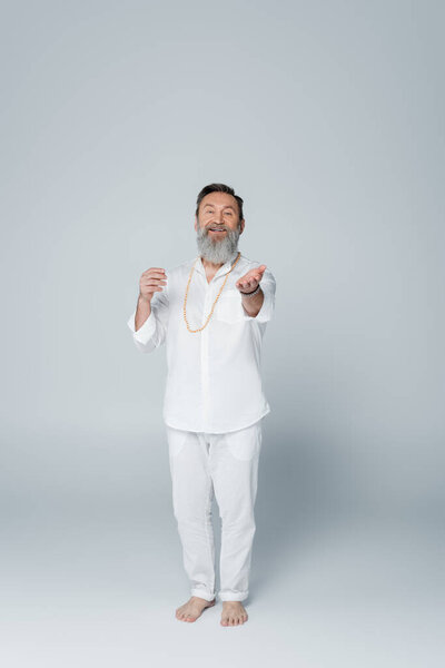 full length of pleased yoga coach in white clothes looking at camera and pointing with hand on grey