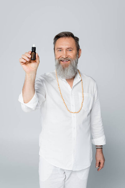 bearded healing guru holding bottle of essential oil and smiling at camera isolated on grey