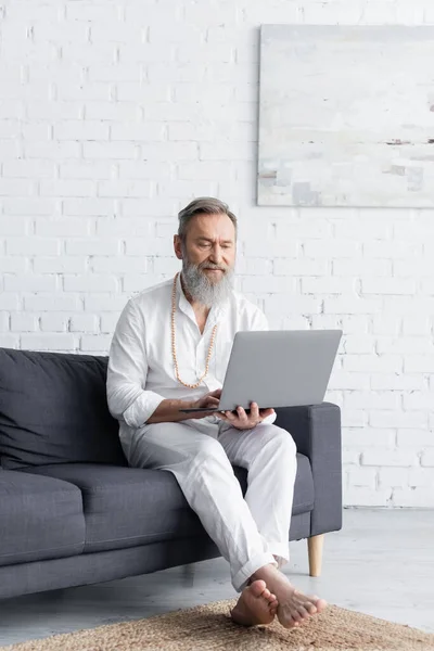 bearded master guru in white clothes sitting with laptop on sofa at home