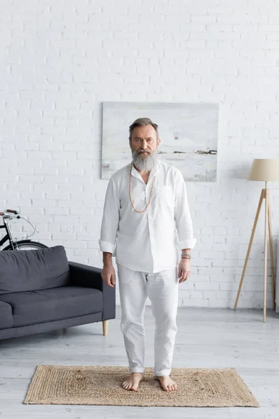 bearded yoga coach in white clothes standing on rug at home and looking at camera