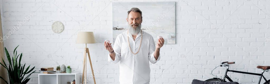 bearded master guru holding selenite crystals and looking at camera in living room, banner