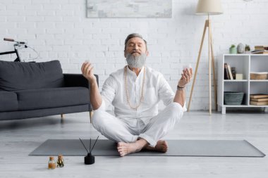 Sakallı yoga ustası aroma çubukları ve kokulu yağların yanında rahat bir şekilde meditasyon yapıyor.