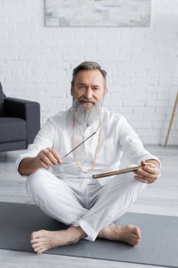 Sakallı yoga öğretmeni elinde aroma çubuklarıyla oturmuş kameraya bakıyor.