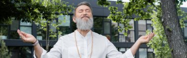 Boncuklar içinde sakallı ruhani koç. Açık havada meditasyon yapıyor, afiş yapıyor.
