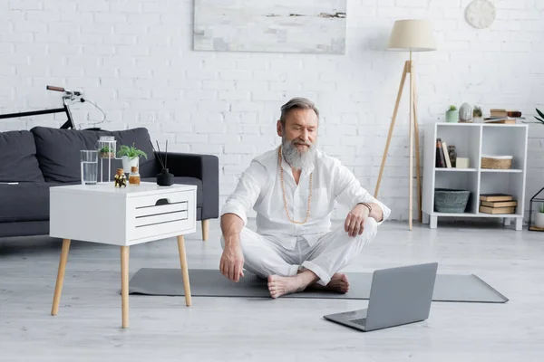 spiritual healing master in easy pose looking at laptop near nightstand with essential oils and aroma sticks