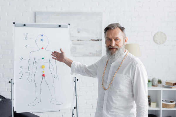 senior ayurveda coach pointing at flip chart with human chakras scheme and looking at camera 