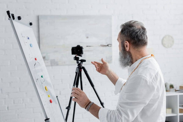 senior healing guru pointing at human chakras system on flip chart near digital camera