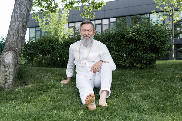 full length of barefoot guru man in white clothes sitting on green grass
