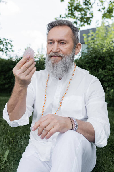 senior master guru in white shirt and beads looking at selenite crystal outdoors