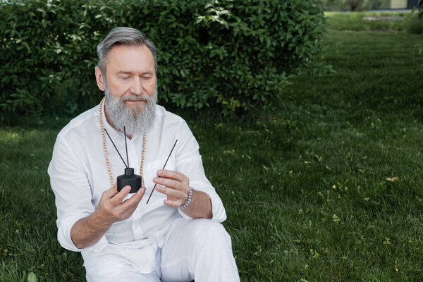 bearded healing guru holding diffuser and aroma sticks while sitting on lawn in garden
