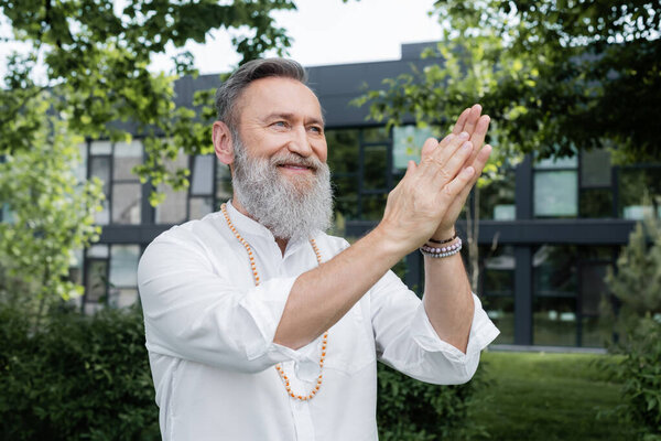 smiling spiritual mentor looking away while meditating with praying hands in park