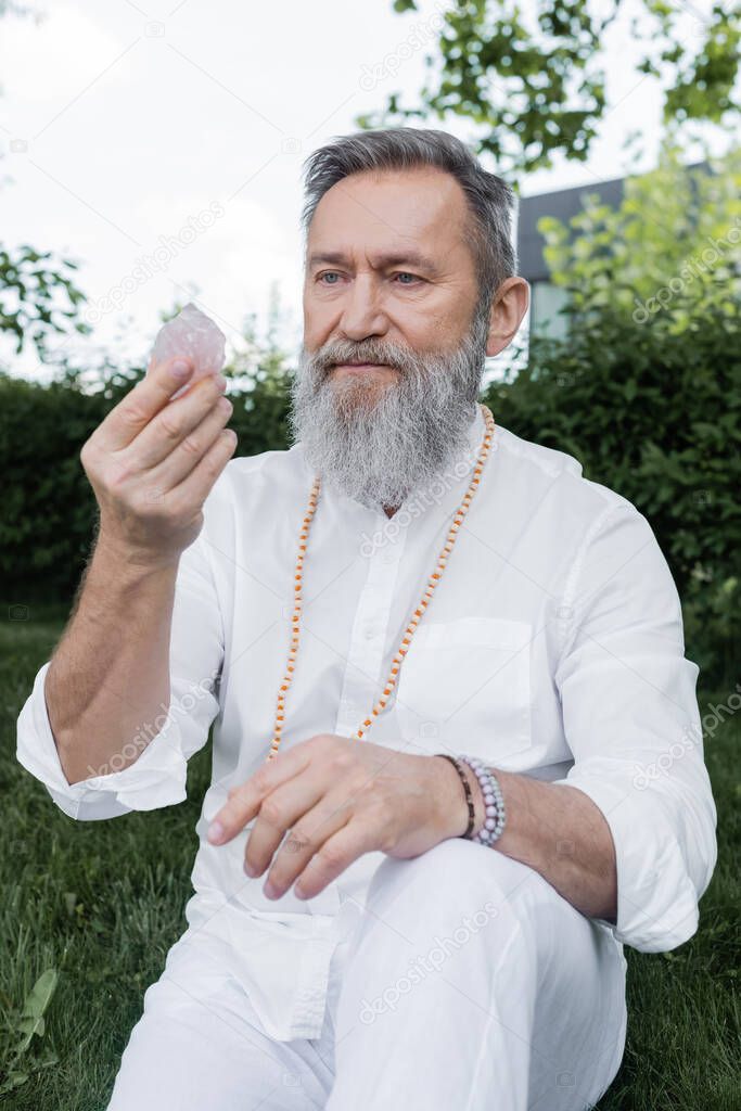 senior master guru in white shirt and beads looking at selenite crystal outdoors