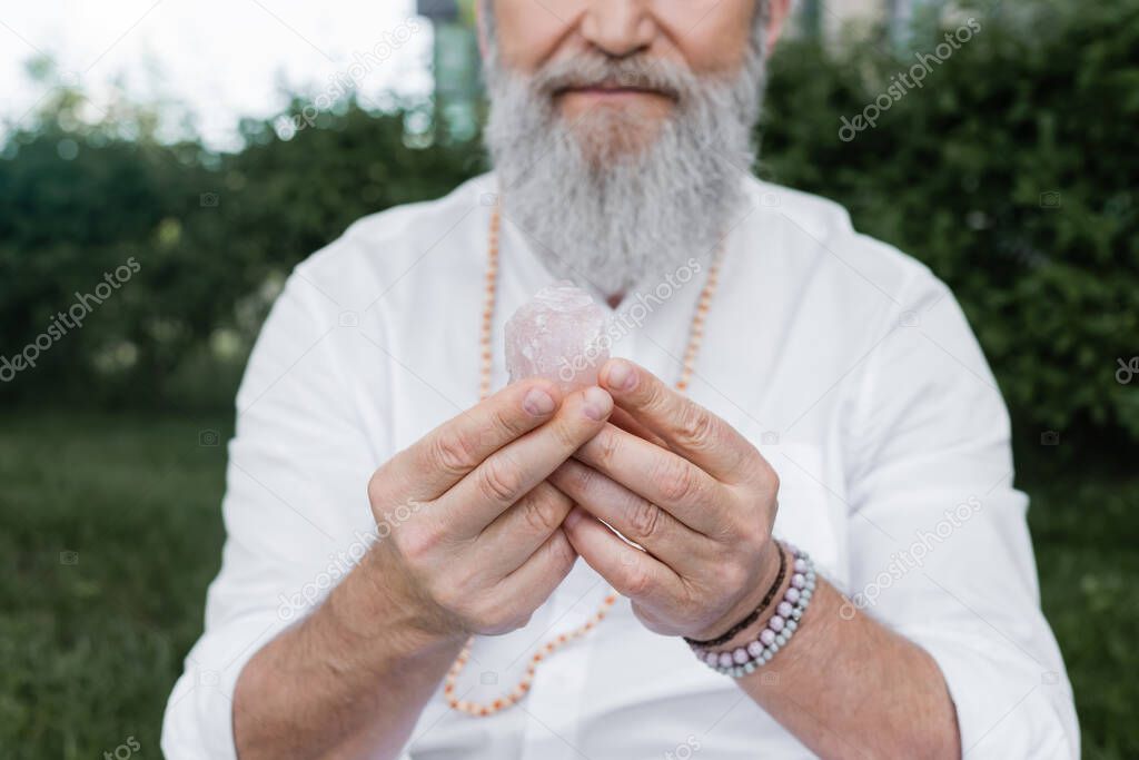 partial view of bearded spiritual mentor in beaded bracelets meditating with selenite stone outdoors