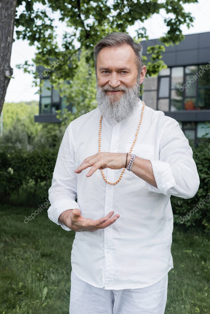 smiling guru man in white shirt showing energy gesture while meditating outdoors
