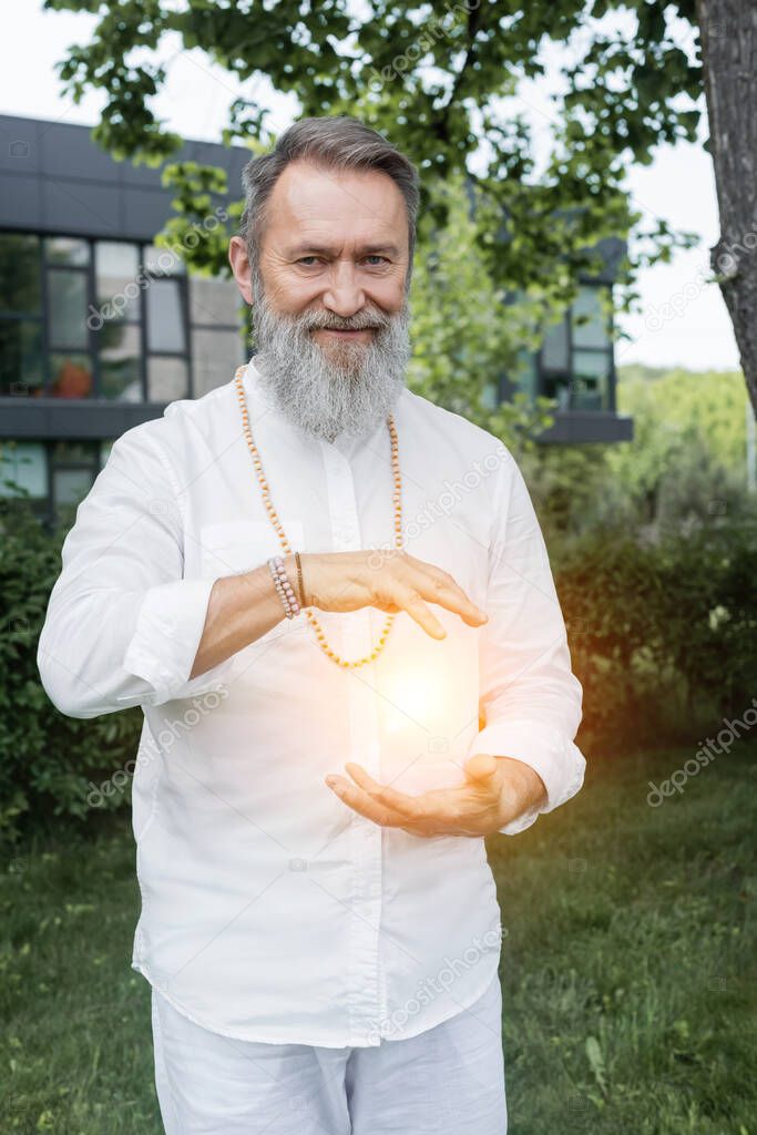 happy guru man looking at camera near shining aura between hands