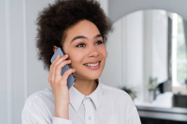 Modern oturma odasında cep telefonuyla konuşan neşeli Afrikalı Amerikalı kadın.