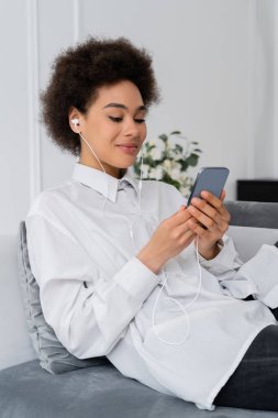 Neşeli Afro-Amerikan kadın, telli kulaklıkla müzik dinliyor ve modern oturma odasında akıllı telefon kullanıyor.