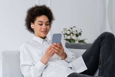 Kıvırcık Afrikalı Amerikalı bir kadın. Kablolu kulaklıkla müzik dinliyor ve modern oturma odasında akıllı telefon kullanıyor.