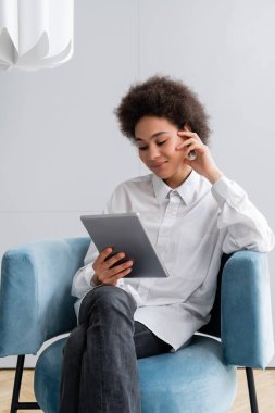 Neşeli Afrikalı Amerikalı kadın mavi kadife koltukta oturuyor ve dijital tablet kullanıyor. 