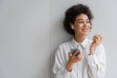 Neşeli Afrikalı Amerikalı kadın kablosuz kulaklıkla müzik dinliyor ve gri duvarın yanında akıllı telefon tutuyor.