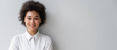Beyaz gömlekli, gri duvarın yanında duran mutlu Afro-Amerikan kadını.