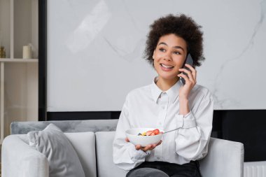 Afro-amerikalı mutlu kadın kahvaltıda kase tutuyor ve akıllı telefondan konuşuyor.