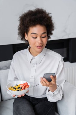 Kıvırcık Afrikalı Amerikalı kadın kahvaltı ve akıllı telefonuyla kase tutuyor.
