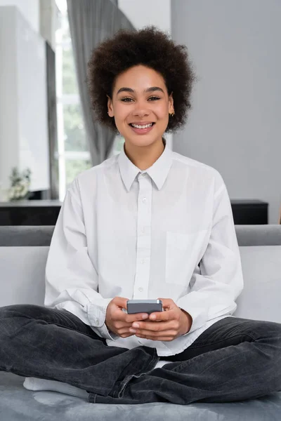 Sonriente Mujer Afroamericana Sosteniendo Teléfono Inteligente Mientras Está Sentado Sofá —  Fotos de Stock