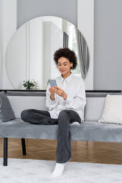pleased african american woman listening music in wired earphones and using smartphone in modern living room