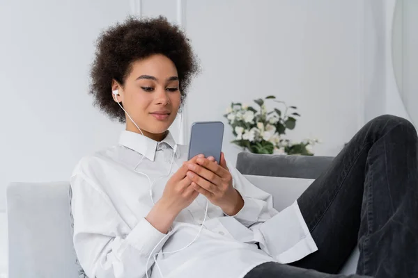 在现代客厅里 卷曲的非洲裔美国女人用有线耳机听音乐 用智能手机 — 图库照片
