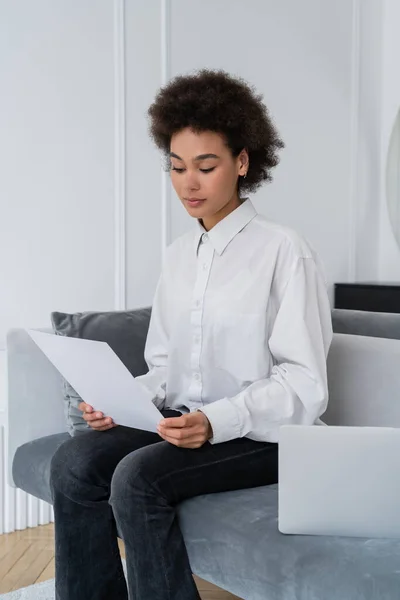 Mujer Afroamericana Mirando Documento Blanco Cerca Computadora Portátil Sofá Gris —  Fotos de Stock