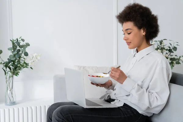 Mujer Afroamericana Sosteniendo Tazón Con Bayas Mientras Película Portátil Sofá — Foto de Stock